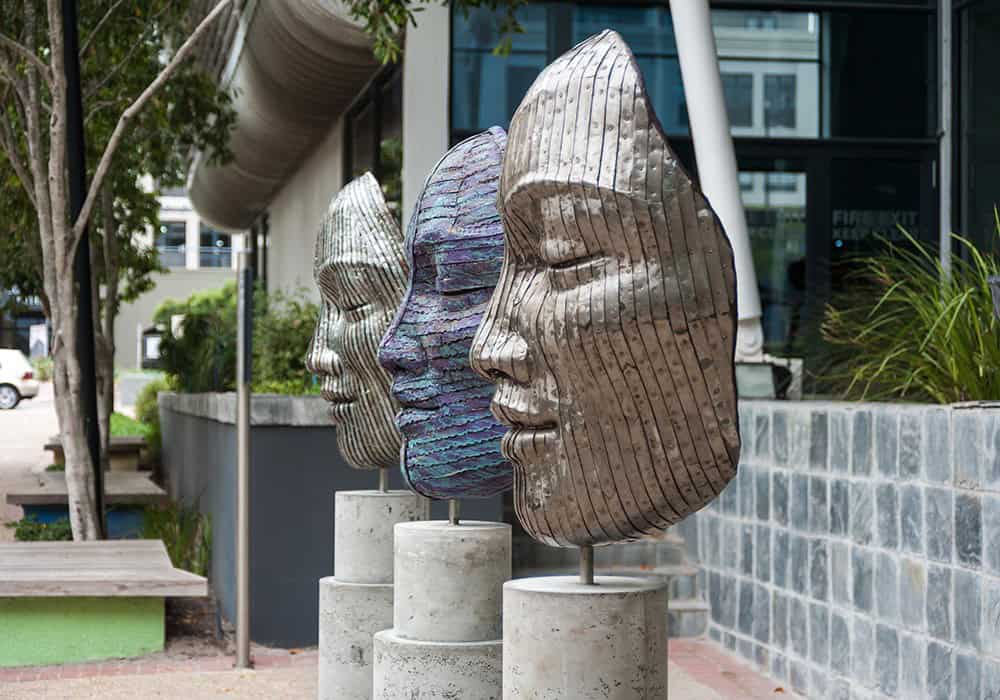 Century City Attractions 2 Three modern metallic face sculptures on concrete pedestals in an outdoor urban setting with greenery.