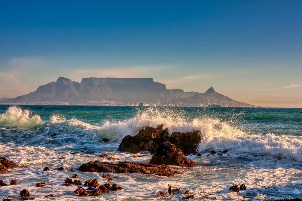 cape-town-usa-destination Stunning view of Table Mountain and Lion's Head in Cape Town, South Africa, seen from across the ocean with waves crashing against coastal rocks in the foreground for Cape Town travel