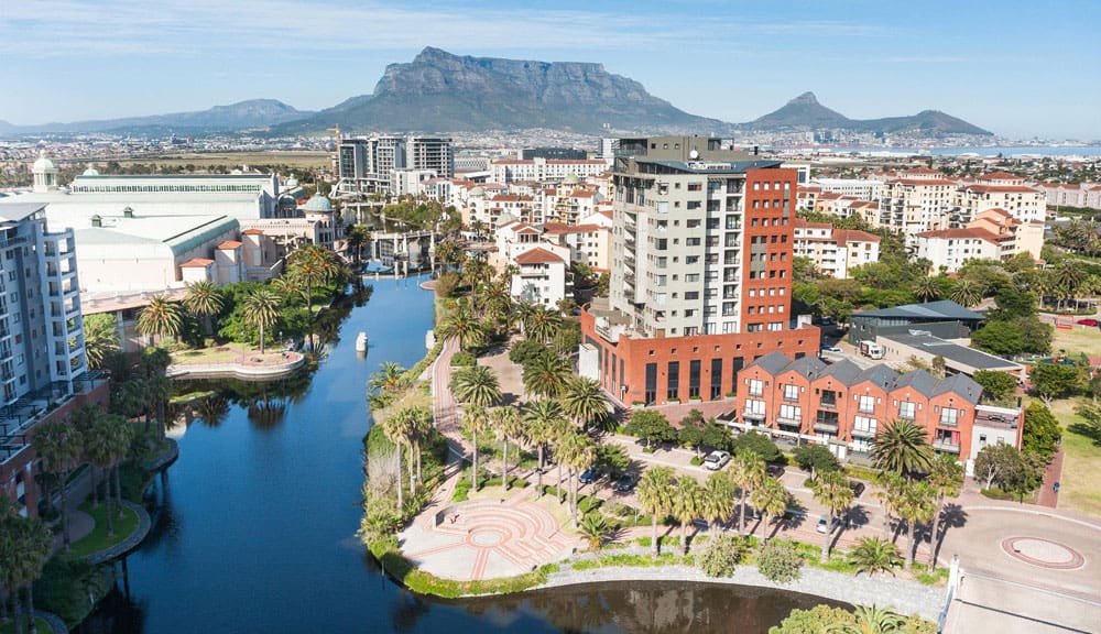 exploring-cape-town-unique-neighbourhoods-2 A river flows through a century city, surrounded by buildings and mountains in the background.