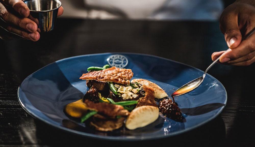 romantic-date-spots-cape-town-2 A chef in Century City holds a spoon above a beautifully plated dish, showcasing culinary artistry and presentation.