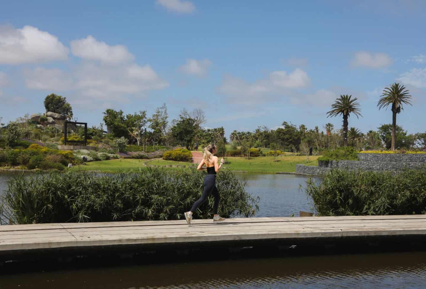hidden-cape-town-hiking-trails to-explore A woman running outdoors, likely engaging in a fitness activity on one of the Cape Town Hiking trails. She is wearing athletic attire, including a black top and dark leggings, and has her hair pulled back. The background features a natural landscape with a body of water